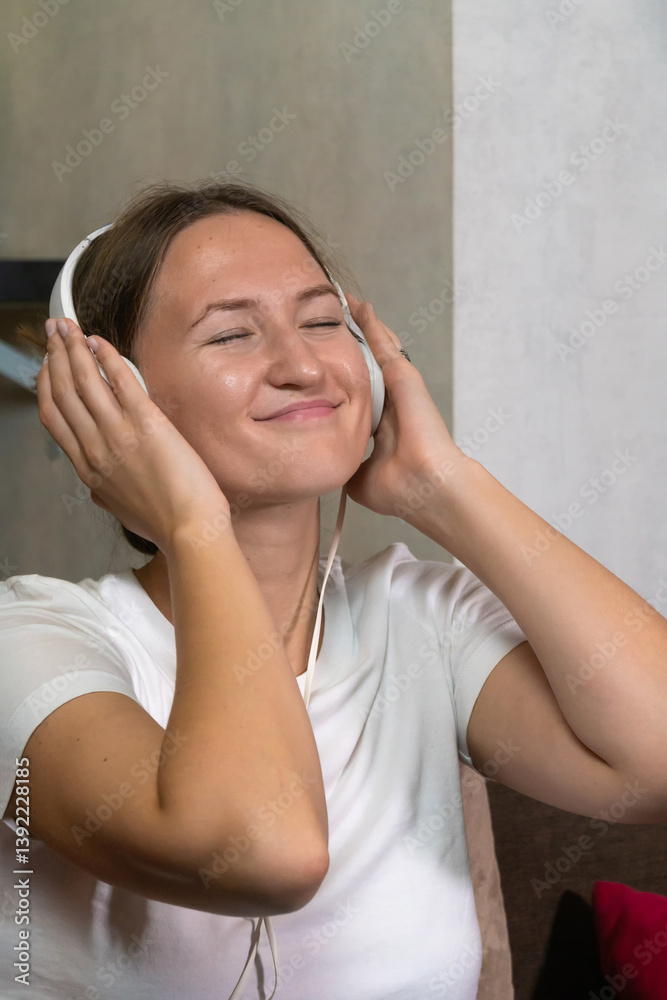 Young happy woman with her eyes closed enjoys music in white headphones while sitting on the couch. Concept of freedom, lifestyle. Vertical photo