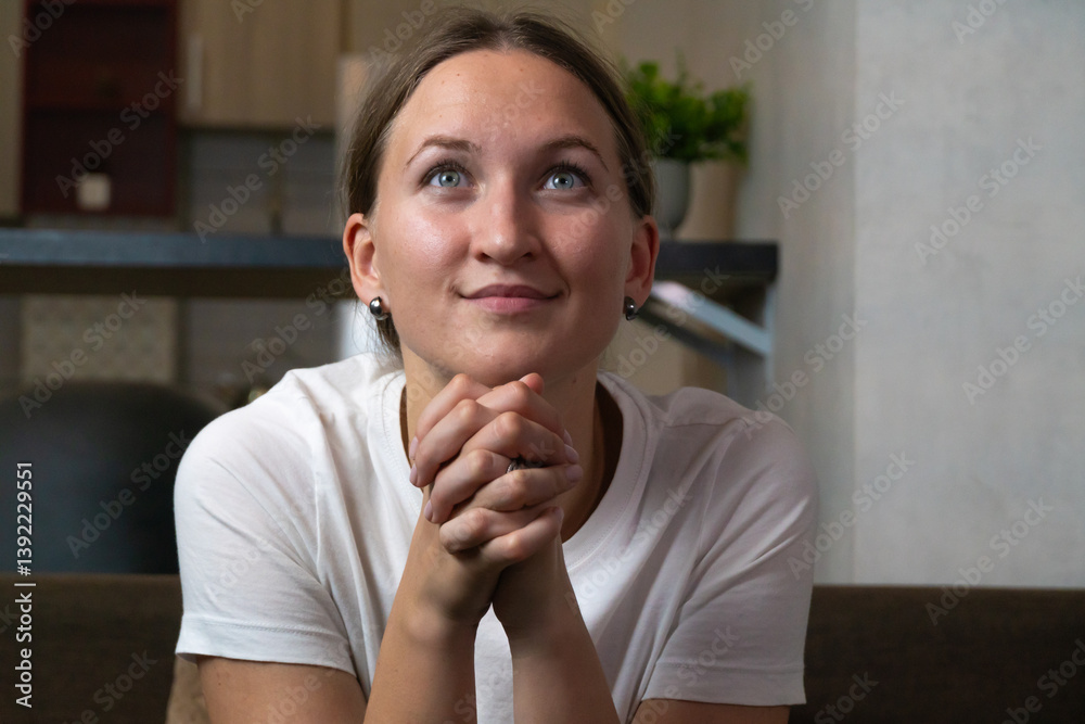 Young woman sitting on a sofa, clasping her hands together in prayer, with a hopeful expression on her face, seeking solace and guidance in her home. Woman is watching TV