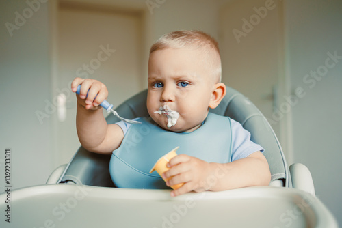 Smiling boy sitting in a high chair, eating yogurt in the kitchen. Healthy food for children. Bio yogurt food for children. Children eating vegetables. Little boy eating dairy products.