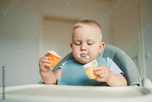 Obraz na płótnie Smiling boy sitting in a high chair, eating yogurt in the kitchen. Healthy food for children. Bio yogurt food for children. Children eating vegetables. Little boy eating dairy products. w salonie