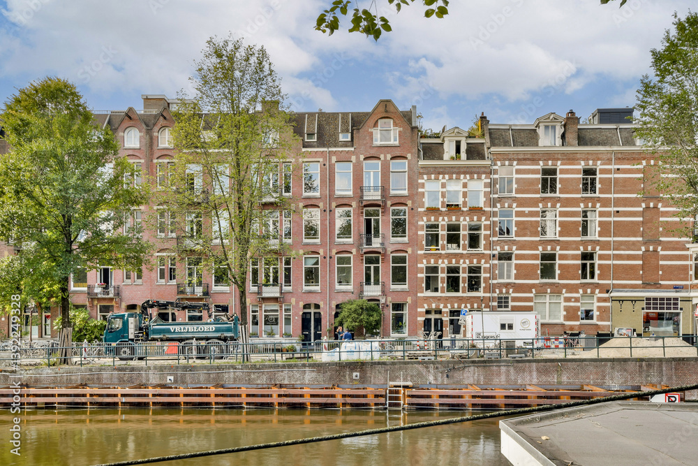 Naklejka premium Picturesque row of historic buildings along a serene canal, exemplifying Amsterdam's unique architectural style with vivid facades and lush greenery.