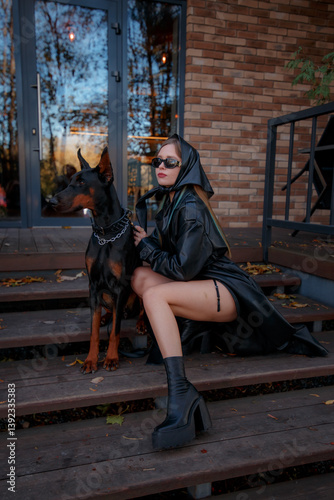 Aesthetic photo of attractive young woman in daring style wearing leather coat and scarf, with doberman dog