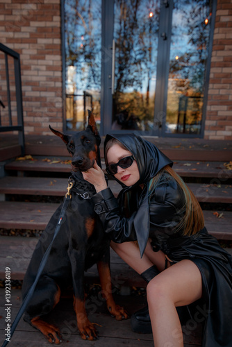 Aesthetic photo of attractive young woman in daring style wearing leather coat and scarf, with doberman dog