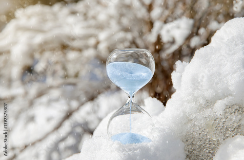 Conceptual image of switching to winter time or fall back, end of daylight saving time. Selective focus on hourglass with blue color sand falling in the winter in snowy nature. photo