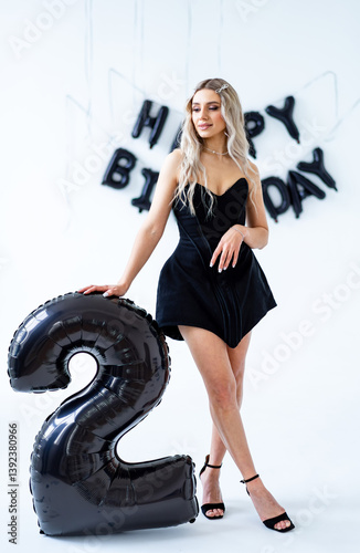 Celebration of second birthday in style. A young adult poses with a large black number two balloon, celebrating a second birthday in a festive setting. photo