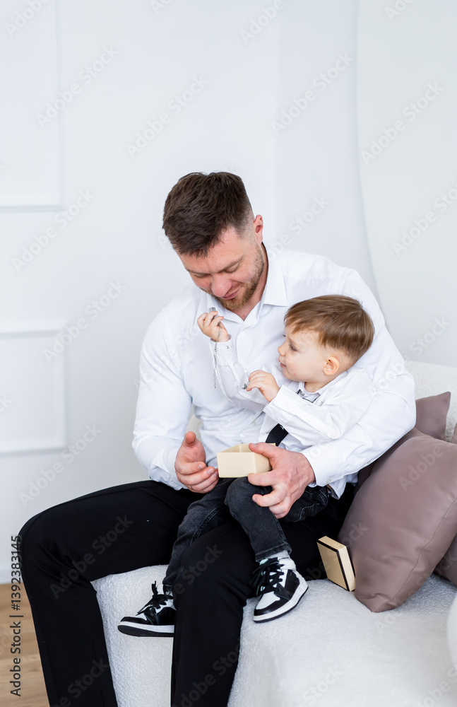 Fototapeta premium Dad and child having fun!. A father sits on a couch, engaging playfully with his young son who is holding a toy.
