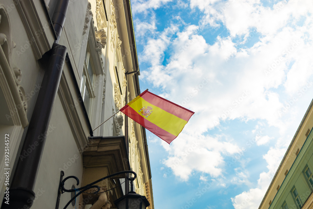 The Spanish flag is proudly waving from a building against a beautiful blue sky with scattered clouds. The flag’s vibrant colors contrast beautifully with the historic architecture, highlighting its
