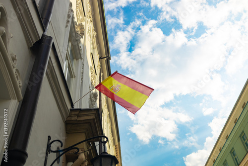 Obraz na płótnie The Spanish flag is proudly waving from a building against a beautiful blue sky with scattered clouds. The flag’s vibrant colors contrast beautifully with the historic architecture, highlighting its w salonie