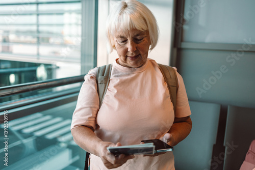 Obraz na płótnie A senior woman smiles while holding her smartphone in the airport terminal. She stands in front of the large glass windows of the terminal, awaiting her flight. The modern airport interior is visible. w salonie