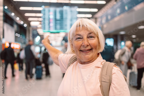 Obraz na płótnie Cheerful elderly woman is pointing at the airport departure board, smiling while surrounded by other travelers. She is excited about her upcoming flight, showcasing the joy of air travel for seniors. w salonie