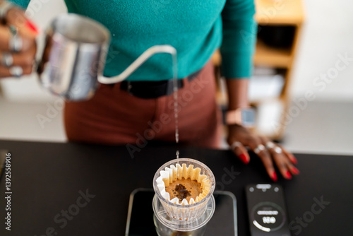A woman pours hot water from a sleek metal kettle into a glass pour-over dripper, slowly wetting the coffee grounds in a careful brewing process in a modern kitchen setting.