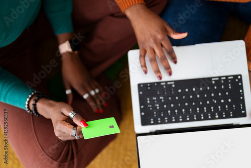 Two women sit on the floor with a laptop, one entering details while the other holds a credit card, making an online purchase in a bright, modern home setting.