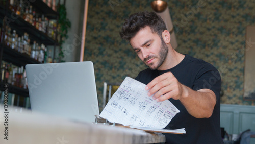 Male Owner Or Manager Of Restaurant Going Through Paperwork With Laptop At Bar