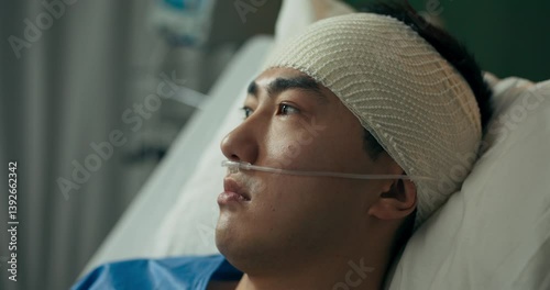 A young man with a bandaged head and an oxygen tube rests in a hospital bed, gazing through the window as he recovers under medical supervision in a quiet and well-equipped ward.