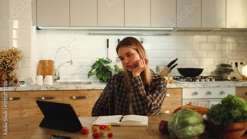 Beautiful girl eating apple watching podcast video on tablet sitting at table in kitchen of her house apartment, recipes, cooking, studying, student, eating, watching media, technology, display