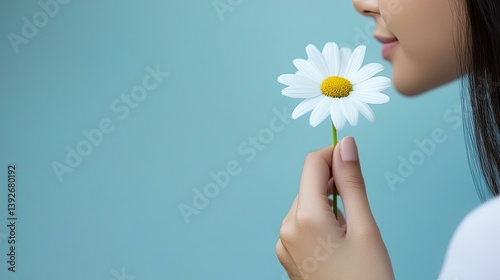 Woman's hand gently holding and smelling a single daisy against a light blue background.