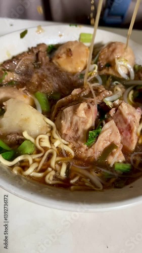 The hand movement of a person stirring meatballs and noodles in a bowl using a spoon to eat.