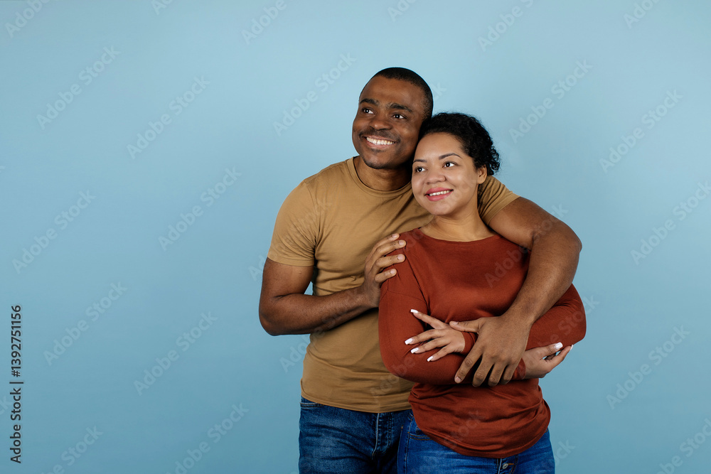 Obraz premium Young black couple embracing affectionately and looking aside at copy space, smiling african american man and woman conveying warmth and emotional closeness, posing on blue studio background