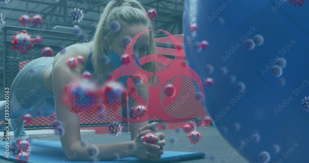 Obraz premium Biohazard symbol and covid-19 cells against woman wearing face mask performing plank exercise at gym