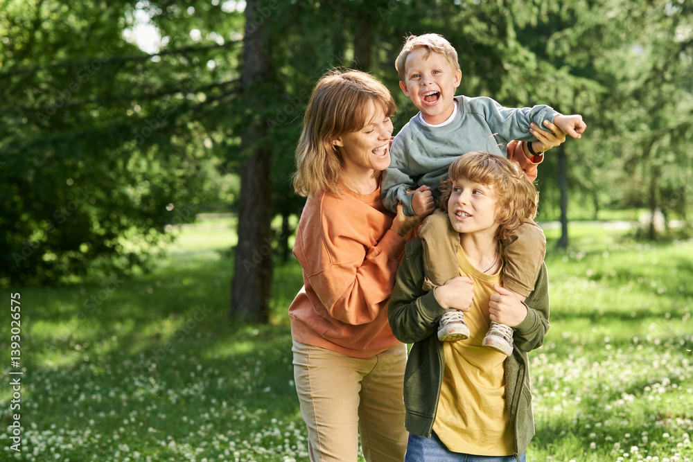 Waist up portrait of young boy holding little brother on shoulders with mother helping family enjoying time together in green park copy space Waist up portrait of young boy holding little brother on shoulders with mother helping family enjoying time together in green park copy space