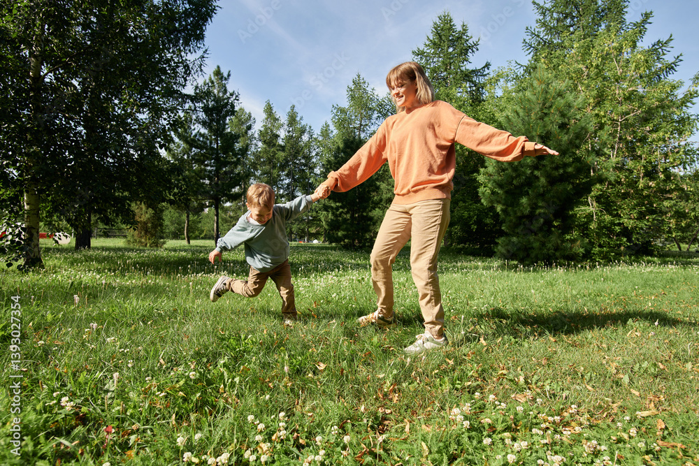 Wide angle view of mother holding hands with little boy while playing on green grass in sunlit park outdoors copy space Wide angle view of mother holding hands with little boy while playing on green grass in sunlit park outdoors copy space