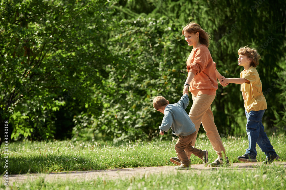 Obraz premium Full length side view of carefree mother with two boys holding hands and walking on path in green forest together lit by sunlight copy space