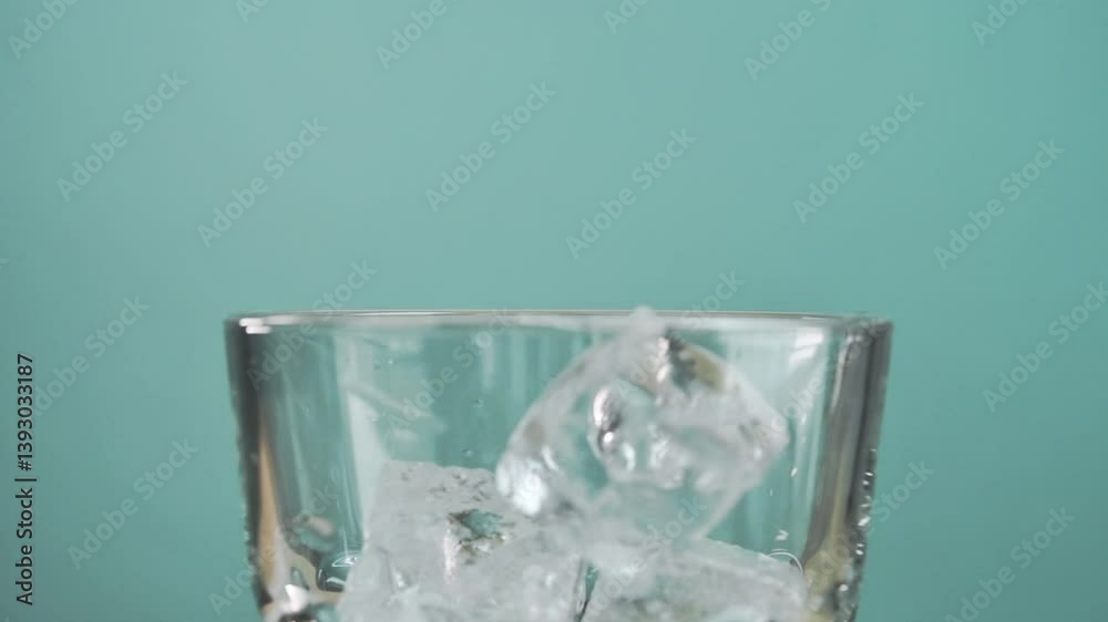 Ice cubes drop into rotating glass on turquoise background, creating a dynamic scene of refreshment, chill and crystal-clear elegance. Ice cubes drop into rotating glass on turquoise background, creating a dynamic scene of refreshment, chill and crystal-clear elegance.