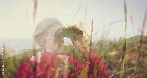 Smiling caucasian senior woman using binoculars in countryside, over moving grasses and sunlight