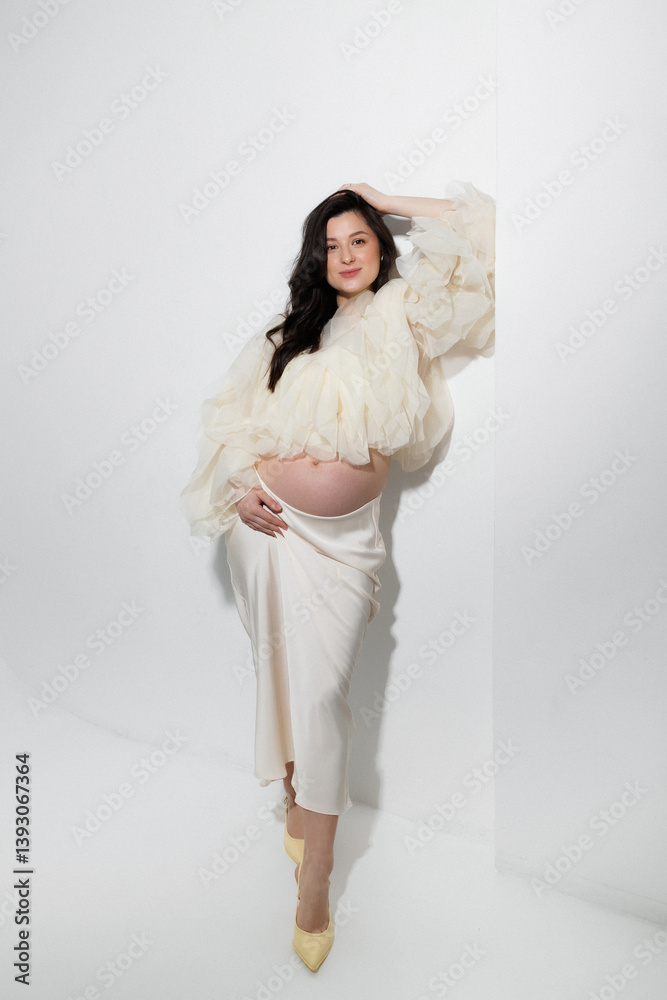 Pregnant woman in a white flowing dress leaning against a white wall. Pregnant woman in a white flowing dress leaning against a white wall.