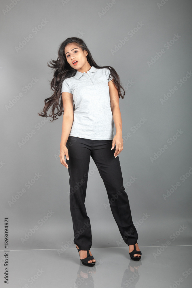 Cheerful young woman dressed in a gray polo t-shirt, black pants, black heels, posing with flowing hair in a studio setting with a grey backdrop. She has a playful expression. Cheerful young woman dressed in a gray polo t-shirt, black pants, black heels, posing with flowing hair in a studio setting with a grey backdrop. She has a playful expression.