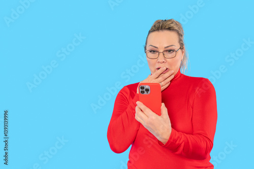 Surprised woman in red top looking at smartphone with astonishment against a bright blue backdrop