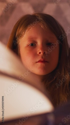 Attentive little girl looks at pages of fairy tale book turned by mother preparing to sleep at gentle light of night lamp in partially illuminated bedroom slow motion
