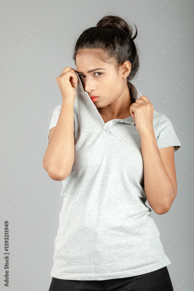 A cheerful corporate office lady or young college student dressed in a gray polo shirt, black pants, black heels, poses in a studio setting with a grey backdrop. She has a playful expression.