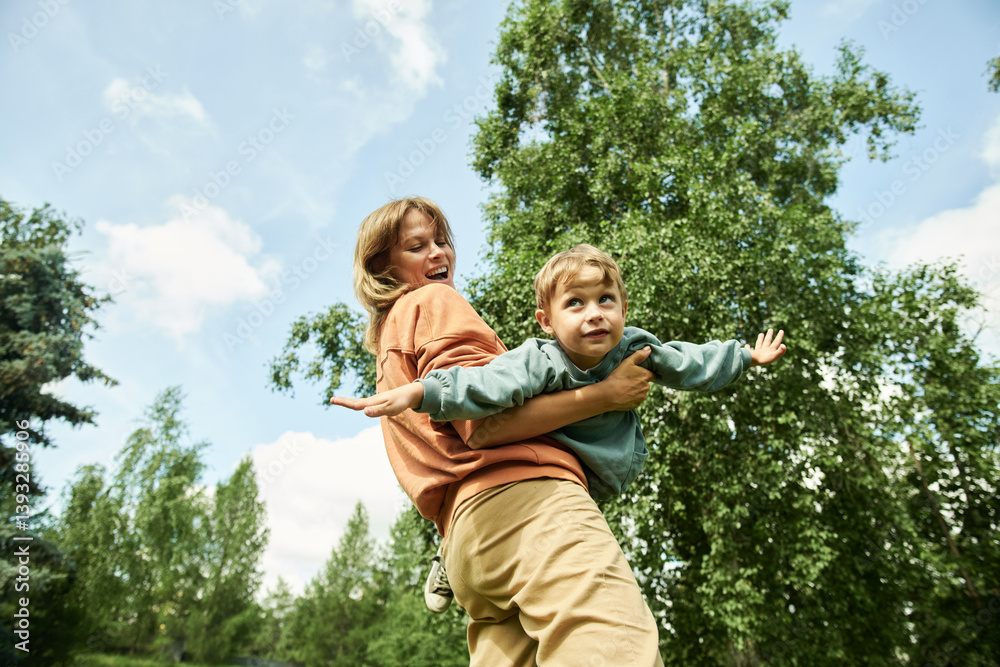 Low angle view at young mother spinning son flying like airplane in green park, enjoying time outdoors Low angle view at young mother spinning son flying like airplane in green park, enjoying time outdoors
