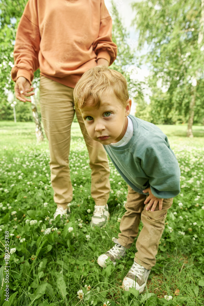 Fototapeta premium Vertical portrait of young boy looking at camera with curiosity standing on green grass with unrecognizable mother in background