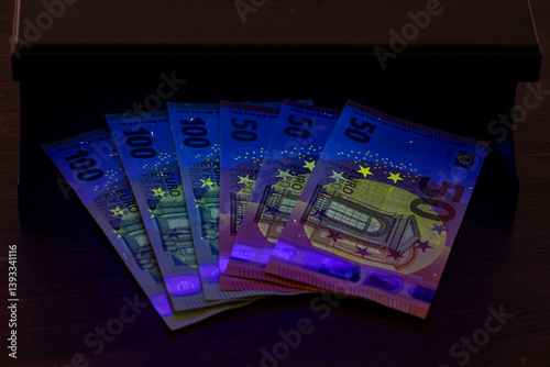 Verifying the authenticity of euro banknotes under ultraviolet light, closeup. Security signs and elements on real 100 and 50 euro banknotes glow under UV light. Checking banknotes for authenticity