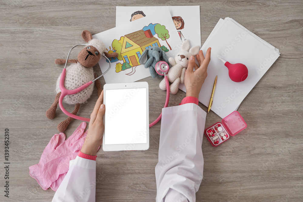 Fototapeta premium Female doctor hands with tablet computer, toys and pills at grey wooden table