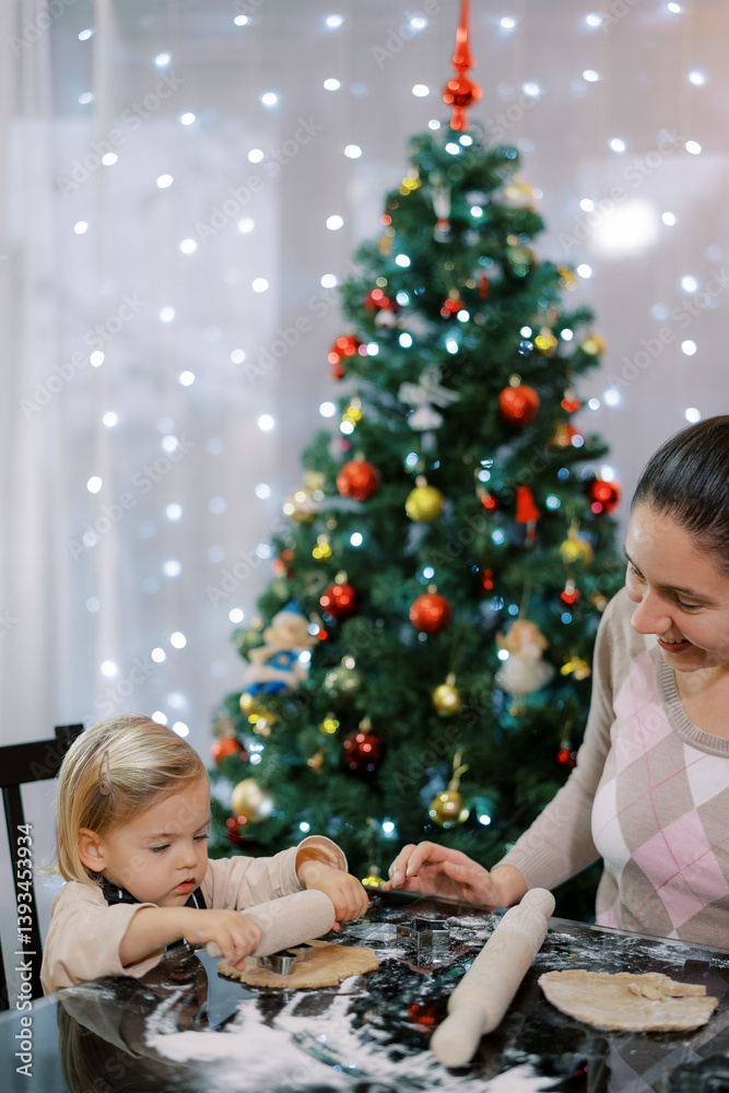 Smiling mother watching little girl rolling dough with small rolling pin on table