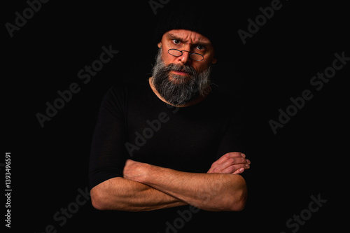 Confident mature man with arms crossed in front of him, wearing a cap and glasses. Staring gaze, gray beard and black vest. Stylish discreet portrait on dark background, low key.