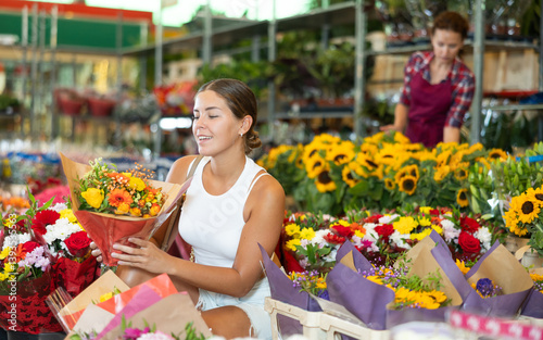 Young woman buyer examines bouquets of fresh flowers, chooses floral arrangement, gift. Girl gets acquainted with assortment of bouquets of cut flowers, examines and chooses.
