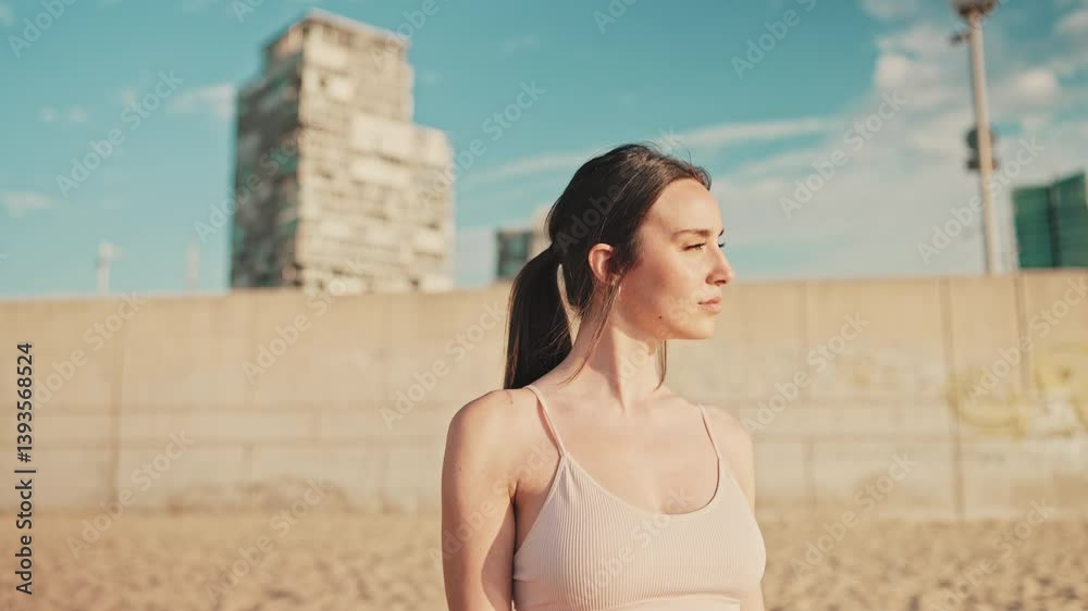 Young athletic woman with long ponytail wearing beige sports top walks along the beach at dawn on modern city background
