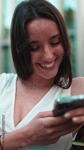 Vertical video, Beautiful woman with freckles and dark loose hair wearing white top sits on bench and watching messages, photos and videos on mobile phone. Camera is getting closer