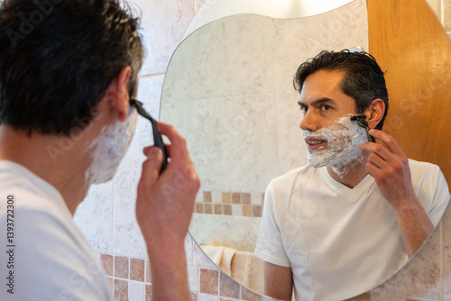 Happy Latino man wearing a white t-shirt shaving his beard in front of the bathroom mirror at home