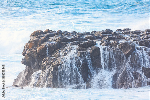 Ocean waves crash against a rugged volcanic rock formation, creating cascading streams of seawater. Dynamic motion captures the raw power and beauty of coastal nature
