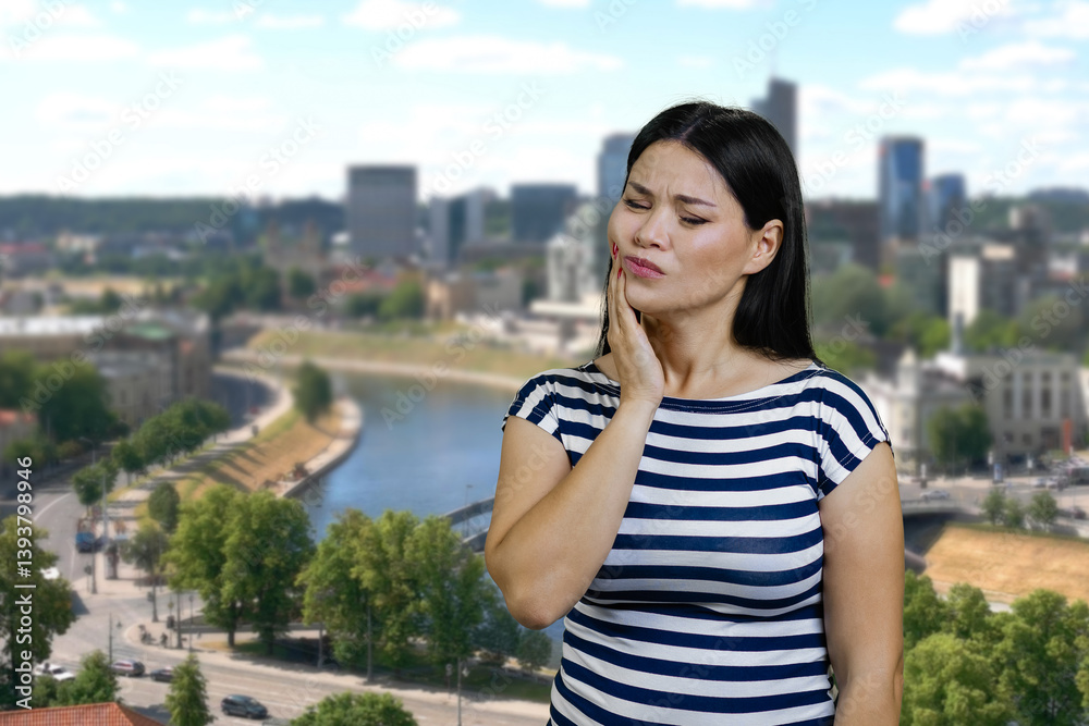 Fototapeta premium Young asian woman touching her cheek because of toothache. Urban cityscape background.