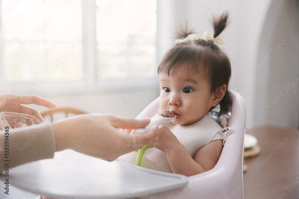 Young Asian child eats while seated in a high chair, lovingly assisted by a parent. A scene representing care and nourishment during early childhood development. Young Asian child eats while seated in a high chair, lovingly assisted by a parent. A scene representing care and nourishment during early childhood development.