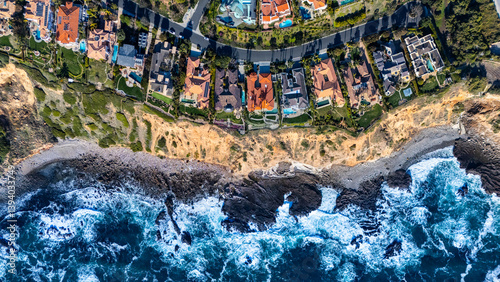 Directly overhead view of upscale homes perched along the rocky shoreline in Rancho Palos Verdes, California. The drone captures the powerful waves of the Pacific Ocean hitting the coast.