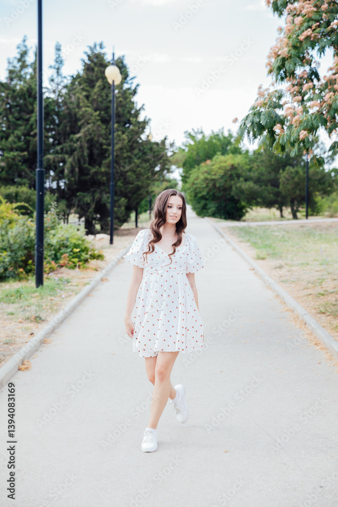 slim fashionable brunette woman in summer white dress walking along the alley in the park slim fashionable brunette woman in summer white dress walking along the alley in the park