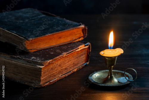 Burning wax candle in bronze candlestick on wooden table next to old books in dark room at night, closeup. Vintage still life with lighting candle near two books