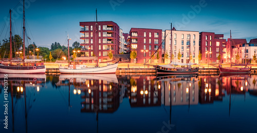 Obraz na płótnie Night summer view of illuminated building and white motorboats on the shore of Ryck river. Amazing evening cityscape of Greifswald town, Germany, Europe. Calm summer scene of Ryck river. w salonie
